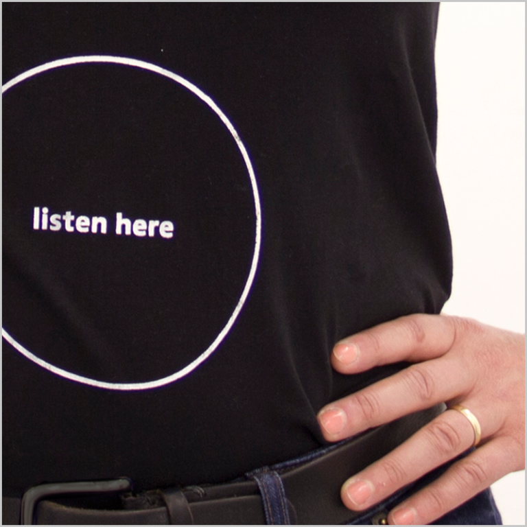 Die Aufschrift "listen here" in einem Kreis ist als weisser Aufdruck auf einem schwarzen T-Shirt